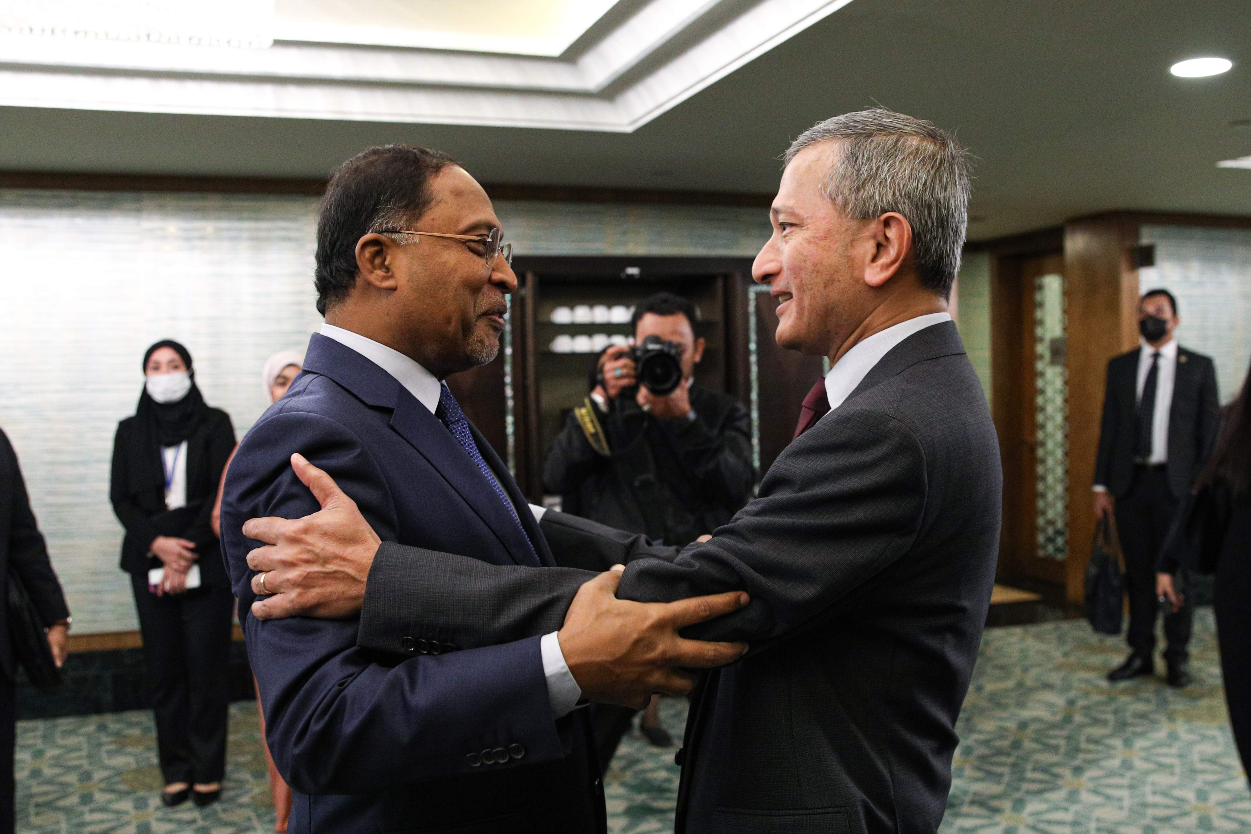 Two men in suits greet, embracing with hands on arms, with others and a photographer in background.